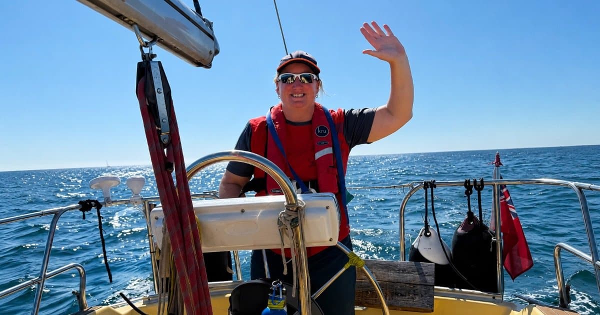 Jenny Whelan at the helm in a red lifejacket, waving, the red ensign flying astern